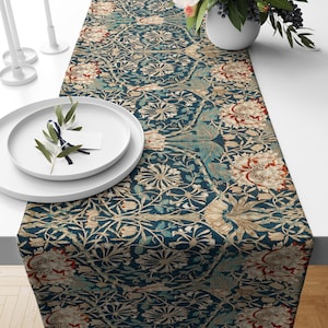 Puede incluir: Un camino de mesa floral azul oscuro con un patrón repetido de flores y follaje estilizados en crema, verde azulado y rojo. El camino de mesa se muestra sobre una mesa blanca, con platos y velas blancas.