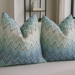 Może przedstawiać: Dwie dekoracyjne poduszki z niebieskim, zielonym i beżowym wzorem zygzaka. Poduszki leżą na jasnoszarej sofie. Poduszki mają miękki, teksturowany wygląd i dodają odrobinę koloru do neutralnej przestrzeni życiowej.