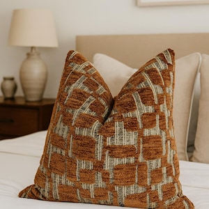 Puede incluir: Un cojín decorativo con un patrón geométrico en tonos marrón, beige y gris. El cojín está colocado sobre una cama blanca, con una lámpara y una mesita de noche en el fondo. El cojín tiene una apariencia texturizada.