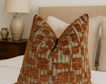 Può includere: Un cuscino decorativo con un motivo geometrico nei toni del marrone, beige e grigio. Il cuscino è posto su un letto bianco, con una lampada e un comodino sullo sfondo. Il cuscino ha un aspetto strutturato.