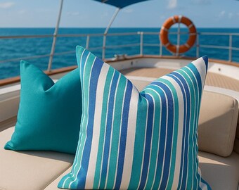 Könnte beinhalten: Zwei dekorative Kissen auf einem beigen Outdoor-Sofa. Ein Kissen ist einfarbig türkis, das andere hat vertikale Streifen in Weiß, Türkis und Blau. Die Kissen befinden sich auf einer Yacht mit dem Meer im Hintergrund.