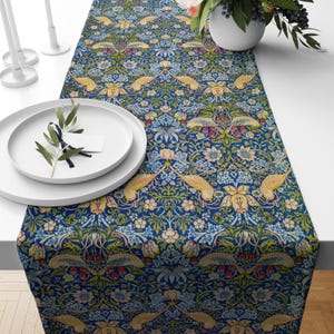 May include: A blue table runner with a floral and bird pattern. The runner features a repeating design of yellow birds, flowers, and leaves in shades of blue, green, and red. The runner is placed on a white table, with white plates and candles.