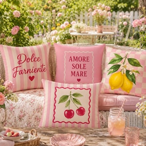 Può includere: Quattro cuscini decorativi con disegni rosa e bianchi. Un cuscino ha strisce verticali e le parole "Dolce Farniente". Un altro ha le parole "Amore Sole Mare". Il terzo cuscino ha un design a ramo di limone e il quarto ha un design a ciliegia.