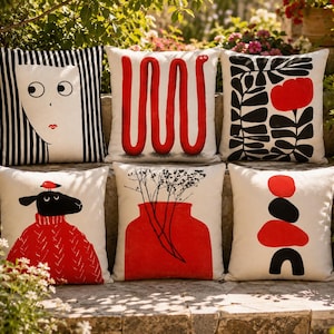 Pode incluir: Seis almofadas decorativas com vários designs. As almofadas apresentam um rosto com listras pretas e brancas, um design vermelho em forma de cobra, padrões florais pretos e vermelhos, uma ovelha com um suéter vermelho e formas abstratas.