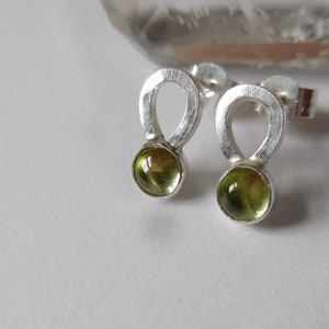 May include: A pair of silver earrings with a green gemstone in the centre of each earring. The earrings are shaped like a figure of eight.