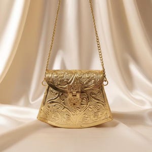 Peut inclure: Un sac à main en métal doré avec un motif floral orné. Le sac a une bandoulière en chaîne et une fermeture à fermoir. Le fond est un tissu doux de couleur crème.