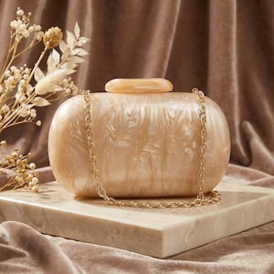 Può includere: Una pochette ovale perlata con una catena dorata. La pochette ha un effetto marmorizzato e una piccola chiusura abbinata. È appoggiata su un blocco di marmo, con fiori secchi sullo sfondo.