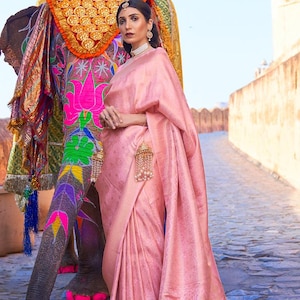 May include: A woman in a pink saree with intricate patterns stands near a decorated elephant. The saree drapes elegantly, showcasing a floral design. The elephant is adorned with colorful decorations, creating a vibrant contrast.