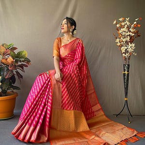 May include: A vibrant pink and orange sari with intricate woven patterns. The sari features a bold striped design in pink, complemented by a gold border and orange tassels. The garment is draped elegantly, showcasing its rich texture and color.