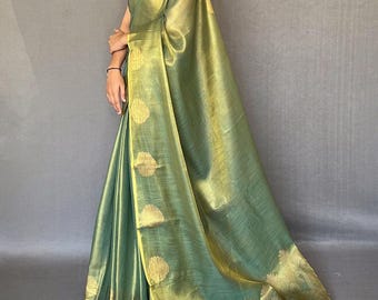 May include: A flowing, sage green saree with gold accents. The fabric has a subtle sheen, and the border features gold circular patterns. The saree is draped elegantly, showcasing its texture and design. The overall look is sophisticated and graceful.