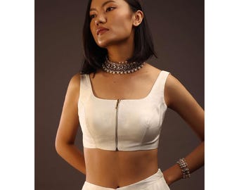May include: A woman wearing a white crop top with a square neckline and a silver zipper. She is also wearing a matching white skirt and a silver necklace and bracelet. The background is a solid, neutral color.