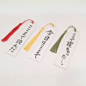 以下が含まれることがあります： 日本語のテキストとカラフルなタッセルが付いた、3つの透明なアクリル製ブックマーク。1つのブックマークには赤いタッセル、別のブックマークには金のタッセル、3つ目には緑のタッセルが付いています。テキストは黒です。
