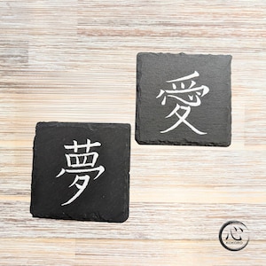 May include: Two square black slate coasters with rough, textured edges. Each coaster features a single white Japanese kanji character engraved in the center. One coaster displays the character for love, and the other displays the character for dream.