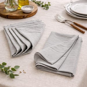 Puede incluir: Dos servilletas de lino gris claro dobladas sobre un mantel beige. Las servilletas están junto a una pila de platos blancos, cubiertos y una pequeña bandeja de madera con una botella de aceite, sal y un jarrón de flores.
