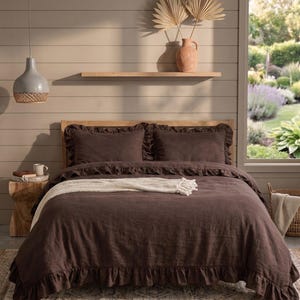 May include: A dark brown bed set with ruffled edges, including a duvet cover and two pillowcases. A cream-colored throw blanket rests on the bed. The bedroom features a wooden bed frame, a small side table, and a decorative vase.
