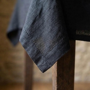 Può includere: Primo piano di un angolo di tovaglia in lino grigio scuro, con un orlo cucito dettagliato. Il tessuto è drappeggiato su un tavolo di legno, creando un'estetica strutturata ed elegante. Ideale per l'arredamento della casa e la ristorazione.