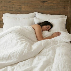 Può includere: Un letto con testiera in legno e biancheria da letto bianca. Una persona sta dormendo sotto le coperte. La biancheria da letto comprende un piumino e cuscini. La stanza ha una finestra con tende.