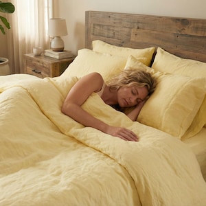Könnte beinhalten: Ein Bett mit gelber Bettwäsche, einschließlich Bettdecke, Kissen und Spannbettlaken. Eine Person schläft in dem Bett. Das Kopfende ist aus Holz. Eine Topfpflanze und eine Lampe sind im Hintergrund zu sehen.