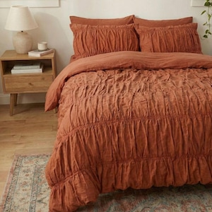 以下が含まれることがあります： くすんだオレンジ色の寝具セット。ギャザーの入った掛け布団カバーと、お揃いの枕カバーが特徴です。寝室には、ランプと柄の入ったラグが置かれた木製のナイトスタンドがあります。全体的に暖かく、居心地の良い雰囲気です。