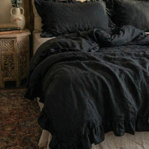 May include: A bed with dark charcoal bedding, including a duvet cover and pillowcases. The bedding has a ruffled edge. The headboard is made of carved wood. A small wooden nightstand and a vase with dried plants are visible.