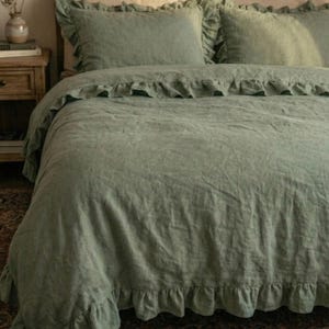 May include: Sage green bedding set with a ruffled duvet cover and pillowcases. The bed is set against a carved wooden headboard. The bedding has a soft, textured appearance, possibly linen. The duvet cover and pillowcases have a frilled edge.