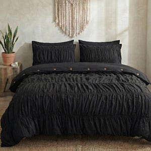 May include: A black bedspread set with two pillowcases. The bedding has a textured, ruched design and wooden button details. The set is displayed on a bed with a woven rug and a macrame wall hanging in the background.