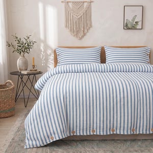 May include: A bedding set featuring a white duvet cover and two matching pillowcases with vertical blue stripes. The duvet cover and pillowcases are accented with small, round, light brown wooden buttons along the edges for a textured detail.