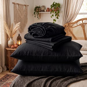 May include: A set of solid black bedding including two rectangular pillows stacked horizontally, a folded flat sheet, and a fitted sheet with elasticized edges, all featuring a matte, textured fabric finish displayed on a bed.