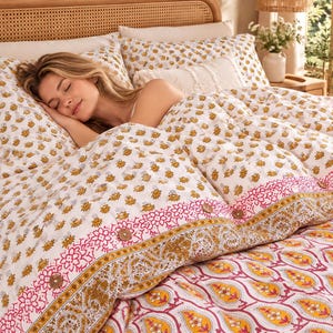 Puede incluir: Una funda nórdica blanca con fundas de almohada a juego que presenta un patrón floral amarillo repetitivo, acentuado por un borde ancho con diseños de cachemira en rosa y amarillo y botones de madera circulares.