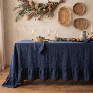 May include: A dark blue rectangular tablecloth with a ruffled edge, draped over a dining table. The table is set with ceramic bowls, wine glasses, a pitcher, and folded napkins, creating a complete dining arrangement for a meal setting.