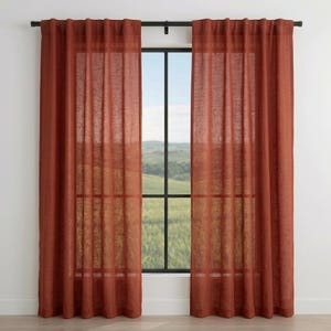 May include: Two rust-colored linen curtains hang on a black rod, framing a window. The curtains are made of a semi-sheer fabric, allowing natural light to filter through. The window reveals a view of a green landscape.