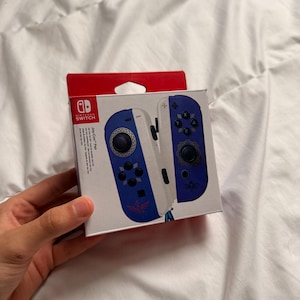May include: A Nintendo Switch Joy-Con Pair box. The box is white with blue and red accents. The Joy-Cons are blue and white with black buttons and a design. The Nintendo Switch logo is visible.