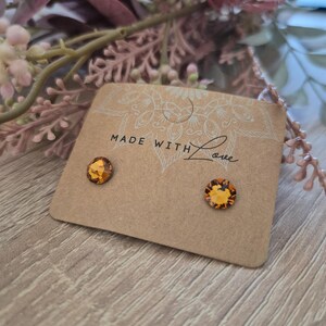 Può includere: Un paio di orecchini a bottone color ambra su una carta beige. La carta ha le parole "MADE WITH Love" stampate sopra. Gli orecchini sono rotondi con un design sfaccettato. Lo sfondo include assi di legno e accenti floreali rosa.