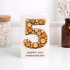 Op de afbeelding: Witte jubileumkaart met een groot houten cijfer 5, gemaakt van kleine houten schijfjes. De tekst "HAPPY 5TH ANNIVERSARY" staat eronder gedrukt. Een glazen pot met witte snoepjes en een bruine vaas staan op de achtergrond.