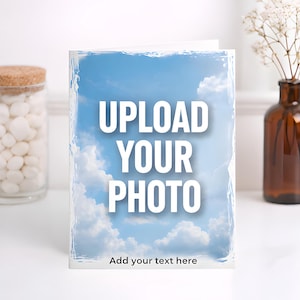 May include: A greeting card with a blue sky and cloud background. The words "UPLOAD YOUR PHOTO" are in large white letters. Below, the text reads "Add your text here". The card is on a white surface.