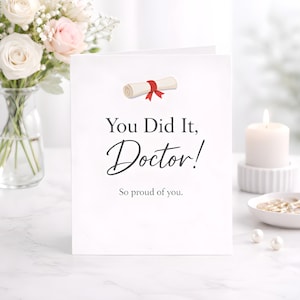 Puede incluir: Tarjeta de graduación blanca con el texto "You Did It, Doctor!" y "So proud of you." Una ilustración de un diploma enrollado con una cinta roja está en la parte superior. Un ramo de rosas blancas y rosas está en el fondo.