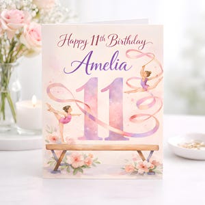 May include: A birthday card with a watercolor design. The card reads "Happy 11th Birthday Amelia" in purple script. The number 11 is large and purple. Gymnasts with ribbons are featured, along with a balance beam and floral accents.
