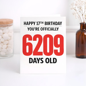May include: A white birthday card with the text "HAPPY 17TH BIRTHDAY YOU'RE OFFICIALLY 6209 DAYS OLD" in black and red. The card is on a white surface, with a jar of white candies and a vase of flowers in the background.