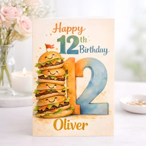 Peut inclure: Carte d'anniversaire avec un design à l'aquarelle. La carte dit "Joyeux 12e anniversaire" avec une pile de hamburgers de dessins animés souriants. Le nom "Oliver" est imprimé au bas de la carte.