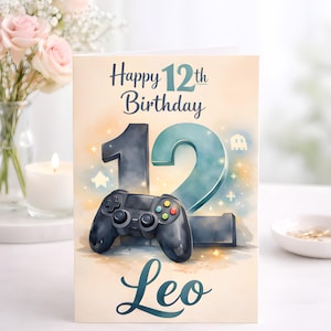 Könnte beinhalten: Geburtstagskarte mit dem Text "Happy 12th Birthday Leo". Die Karte zeigt ein Aquarelldesign mit einem schwarzen Gamecontroller und der Zahl 12 in Blau. Eine kleine Geistergrafik ist ebenfalls sichtbar.