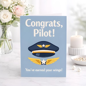May include: A light blue greeting card with the text "Congrats, Pilot!" and "You've earned your wings!" features a navy blue pilot's hat and gold wings. A bouquet of flowers and a lit candle are in the background.