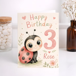 Peut inclure: Une carte d'anniversaire avec une illustration aquarelle d'une coccinelle aux grands yeux, le texte Happy Birthday, un grand chiffre 3 rose à pois et le prénom Rose en écriture cursive rose, agrémentée de petits cœurs et de fleurs.
