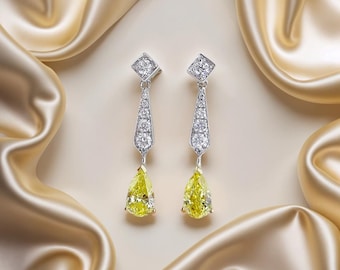 May include: A pair of elegant drop earrings. Each earring features a square-cut diamond at the top, a tapered section with multiple small diamonds, and a large, pear-shaped yellow diamond at the bottom. The earrings are set in silver or white gold and are displayed against a backdrop of draped, golden fabric.