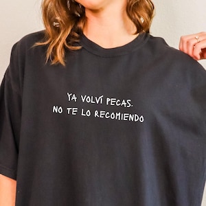 May include: Black oversized t-shirt with white text that reads "YA VOLVÍ PECAS. NO TE LO RECOMIENDO." The shirt is a dark charcoal color and has short sleeves. The model is wearing the shirt.