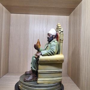 May include: A detailed figurine of a bearded person wearing a white turban and a green military-style jacket, seated on an ornate gold-coloured throne. The figure holds a small, translucent amber-coloured object in one hand and wears brown boots.