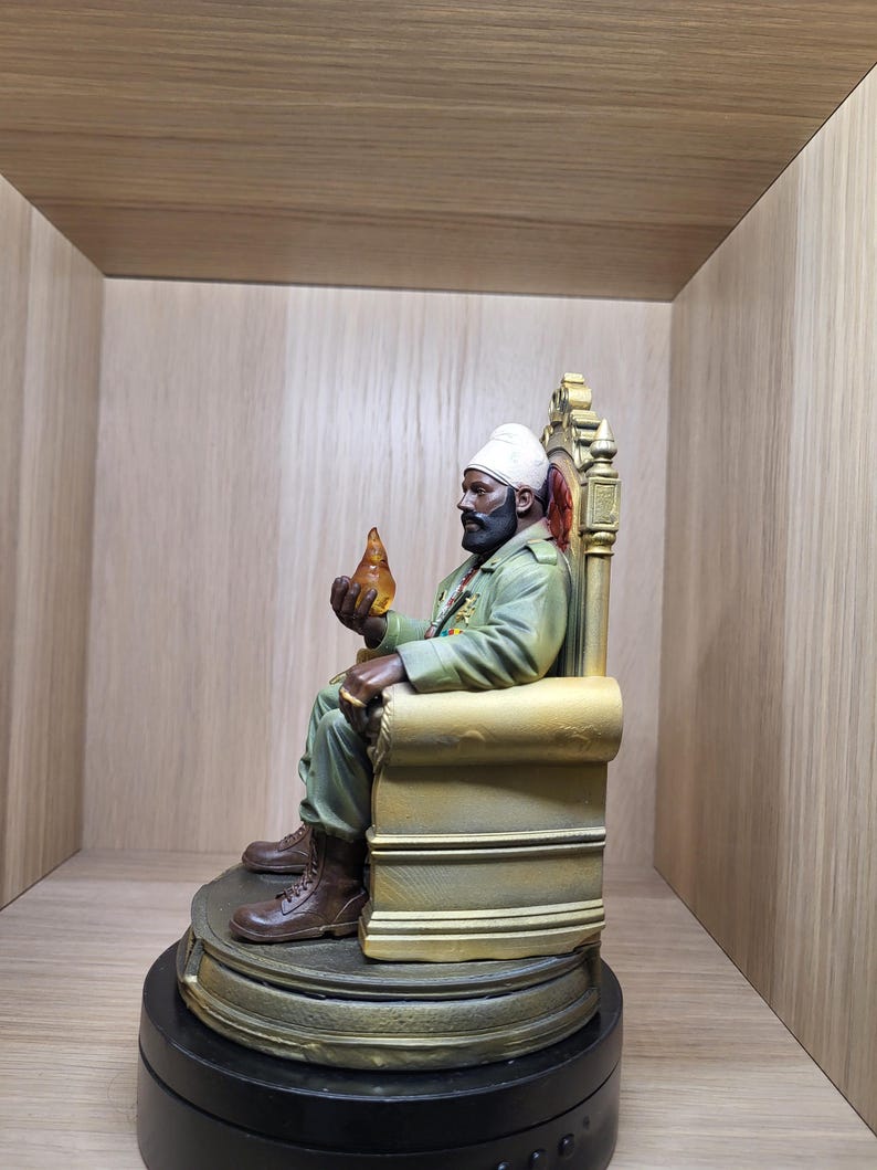 May include: A detailed figurine of a bearded person wearing a white turban and a green military-style jacket, seated on an ornate gold-coloured throne. The figure holds a small, translucent amber-coloured object in one hand and wears brown boots.