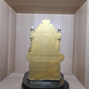 May include: A gold-coloured statue of a seated figure on a throne, viewed from the back. The throne features a tall, ornate backrest with a crown-like top, pointed finials on the sides, and a wide base resting on a circular black stand.