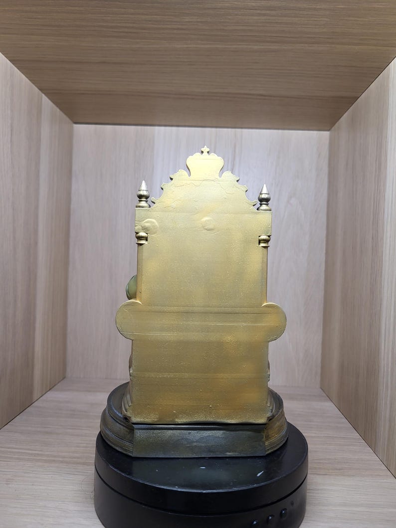 May include: A gold-coloured statue of a seated figure on a throne, viewed from the back. The throne features a tall, ornate backrest with a crown-like top, pointed finials on the sides, and a wide base resting on a circular black stand.