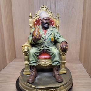 May include: A detailed figurine of a bearded figure wearing a light green military-style jacket and a head wrap, seated on an ornate gold throne with red upholstery. The figure holds a translucent orange flame and has the name CAPLETON on the base.