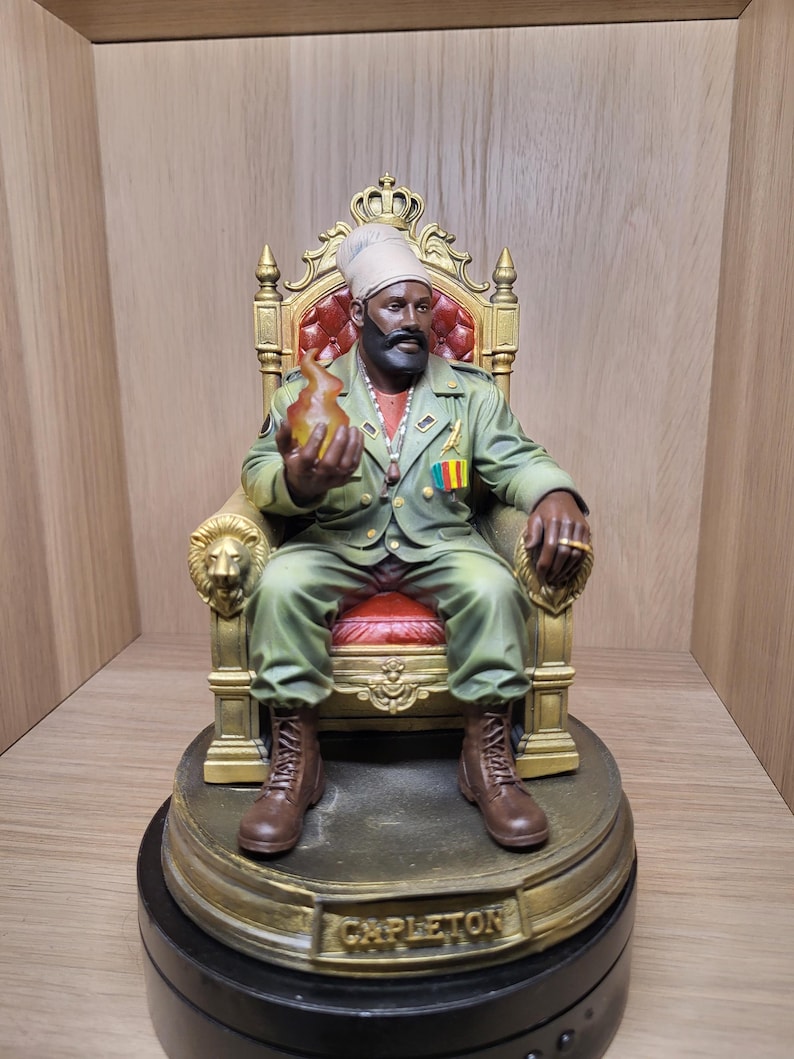 May include: A detailed figurine of a bearded figure wearing a light green military-style jacket and a head wrap, seated on an ornate gold throne with red upholstery. The figure holds a translucent orange flame and has the name CAPLETON on the base.